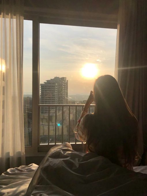 The image shows a woman sitting on her bed with a view of a city skyline outside her window, looking at the sunset. The scene suggests that she might be enjoying some leisure time or contemplating something while taking in the beautiful view. The photo is captioned "Hey friends I just joined @RiverdotInc a super easy way to earn River Points. No big investment needed ... these points will turn into $RIVER tokens later". This implies that the woman has recently joined an account on the platform and is using it to earn River Points, which can be converted into tokens for various purposes.