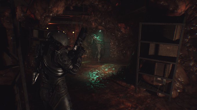 A dark video game screenshot showing a character in a black suit holding a gun, aiming at another character standing near metal shelving in a fleshy, dimly lit corridor.