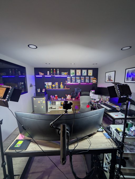 A content creator's streaming setup featuring a wide curved monitor, professional lighting panels, a camera on a tripod, and shelves displaying various figurines and memorabilia.