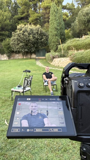 A behind-the-scenes view of a camera operator filming a man seated outdoors in a garden setting, with the subject visible on the camera's monitor.