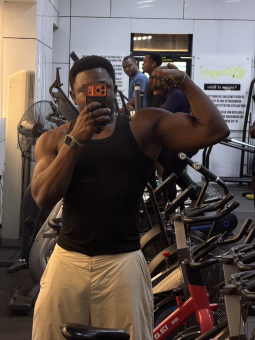 A man takes a mirror selfie at the gym, flexing his right bicep while wearing a black tank top and white shorts, with stationary bikes and a "hagurQ" sign visible in the background. The post's mention of "Back from the gym" aligns perfectly with the image's gym setting, while "What does the market have for us today" suggests he's likely a trader checking in after his workout.