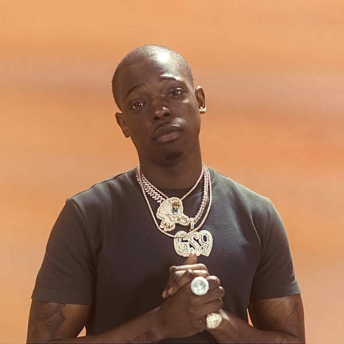 A man wearing a black t-shirt and large diamond-encrusted pendant chains spelling 'ESQ' poses against a brown backdrop.