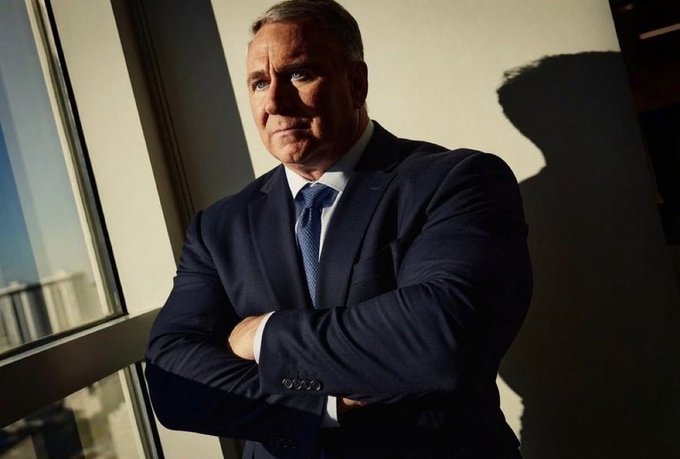 A portrait of a middle-aged man in a navy blue suit and tie, standing with his arms crossed and leaning against a wall in sunlight.