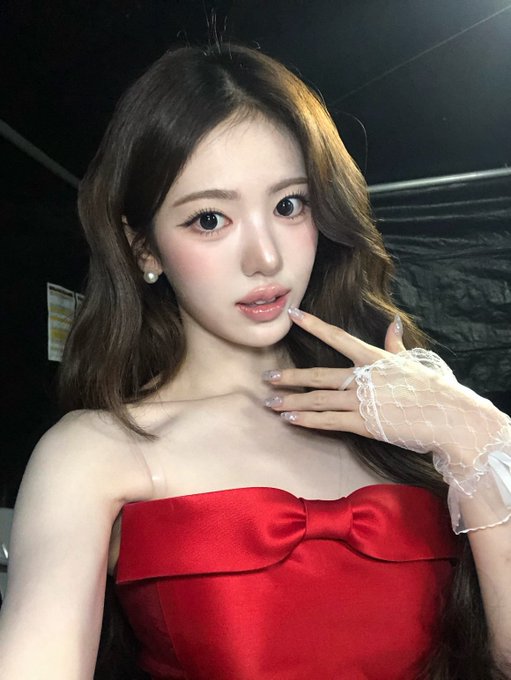 A young woman with long brown hair wears a red strapless top with a bow detail and a white lace fingerless glove, posing with her hand near her face.