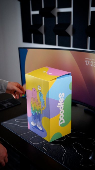 A colorful 'Doodles' branded box with a rainbow character design sitting on a desk mat next to a computer monitor, with a child's hand reaching toward it.