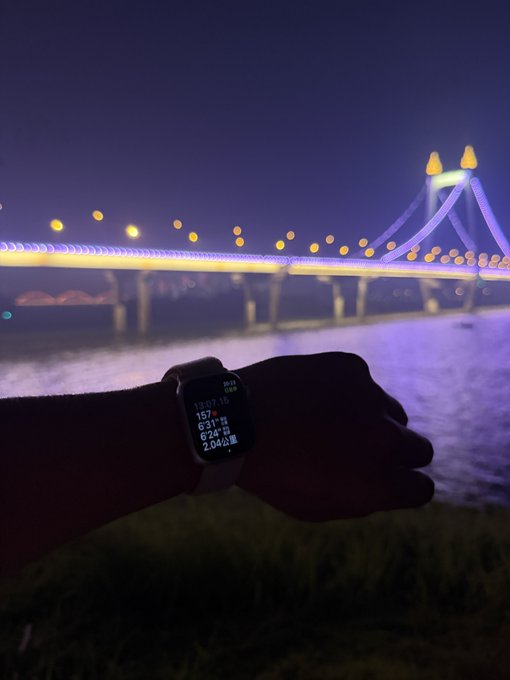 A nighttime photograph of a wrist wearing a smartwatch displaying workout metrics, with an illuminated bridge in the background.