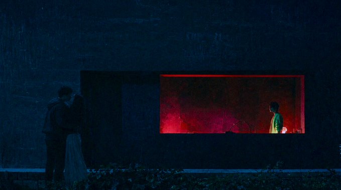 A cinematic, dark scene showing a couple kissing in the rain outside a dimly lit building, while a person in a green shirt stands inside near a red-lit window.