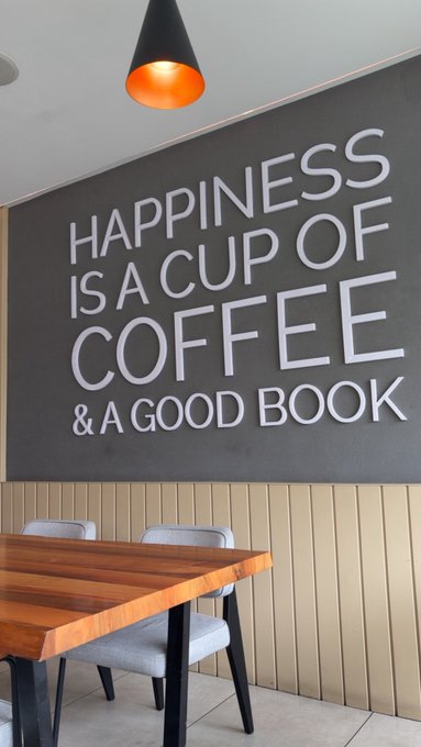A modern café space featuring wall text that reads "HAPPINESS IS A CUP OF COFFEE & A GOOD BOOK," accompanied by a wooden table, gray upholstered chairs, and a black pendant light with a warm copper interior—setting the perfect morning scene for a "Gm" greeting.