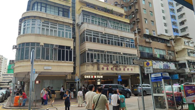 A street corner in Hong Kong featuring older residential buildings with shops on the ground floor, including signs for 'CHAIR' and 'ONE D M SUM'.