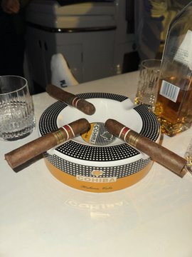 An ashtray designed like a baseball with three lit cigars resting on it, accompanied by a glass tumbler and a bottle of whiskey, sitting on a table in what appears to be a bar or lounge setting.