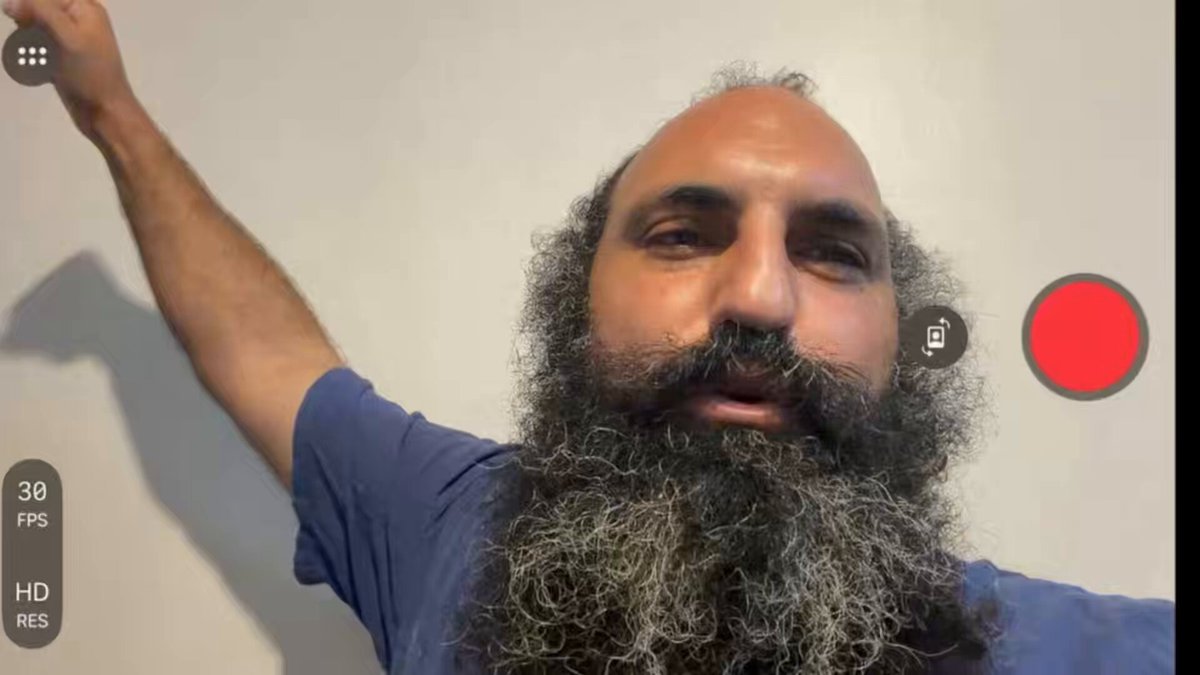 The image shows a bearded man with a red dot on his forehead, likely indicating he is participating in an online meeting or event. The background of the photo has a beige wall and a white door. The caption reads "BITCOIN 7% DROP: I LOCKED IN 70% OF tech stock profits $BTC ... GOOG". This could suggest that the man is engaged in financial discussions related to technology companies such as Bitcoin or some other stock market activity, possibly during an online class or webinar where he can lock in his participation.