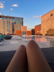 A first-person view of someone relaxing by a rooftop pool, with their legs visible in the foreground and modern apartment buildings and a clear blue sky in the background.