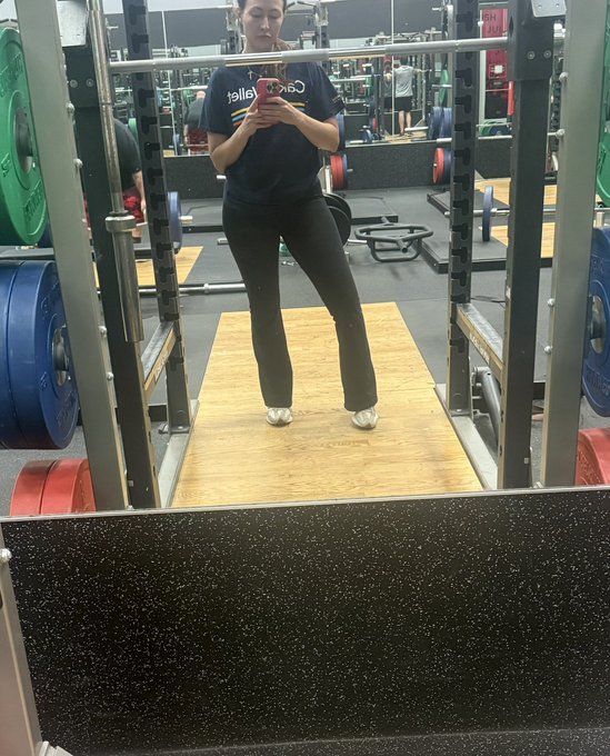 A mirror selfie taken in a gym shows a woman wearing a black 'Coca-Cola' t-shirt and gray flared leggings, standing between weight racks with a smartphone.