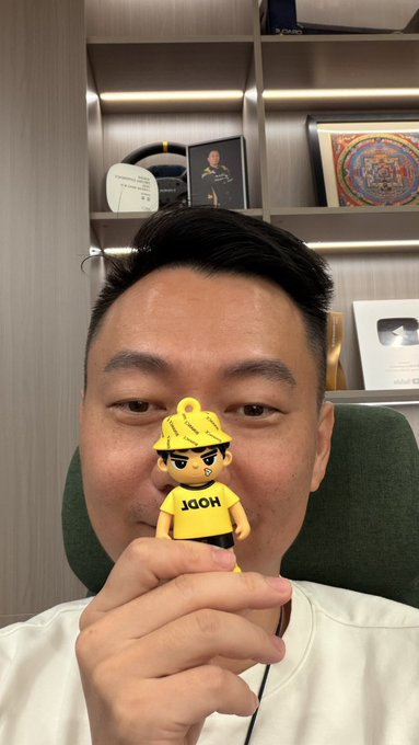 The image features a man wearing a yellow shirt with black text, holding up a small figurine of a person or character that says "jojo" on their shirt. The man appears to be posing for a photo while holding the figurine. The background includes bookshelves and other items, suggesting an indoor setting like a home office or a personal space.