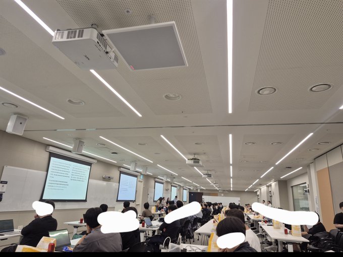 A photograph of a university lecture hall with many students seated at desks, facing projector screens displaying presentation content, with modern ceiling lighting and whiteboards visible, taken from the back of the room.