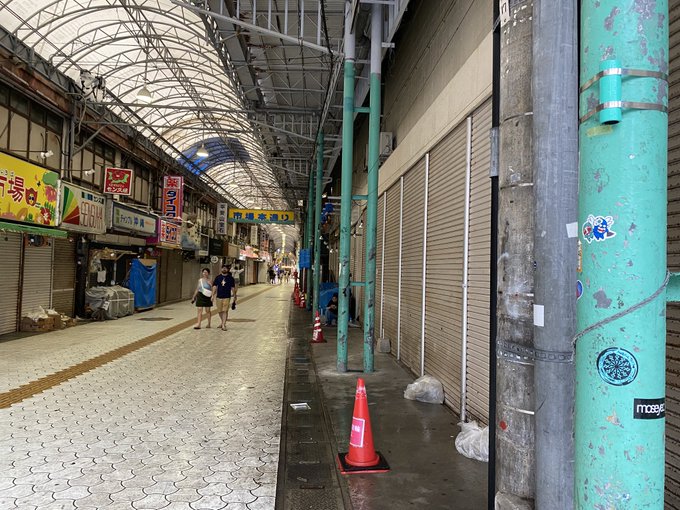 A covered shopping alley in Japan appears largely closed, with metal shutters pulled down along most storefronts and only a few people walking down the tiled walkway past a bright orange traffic cone and weathered teal support pillars—matching the quiet, off-hours atmosphere of "the Alley @Azuki" referenced in the post.