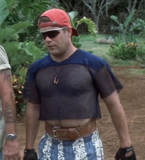 A man wearing an orange backwards cap, sunglasses, a mesh crop top, and a large brown belt, standing outdoors in a tropical setting.