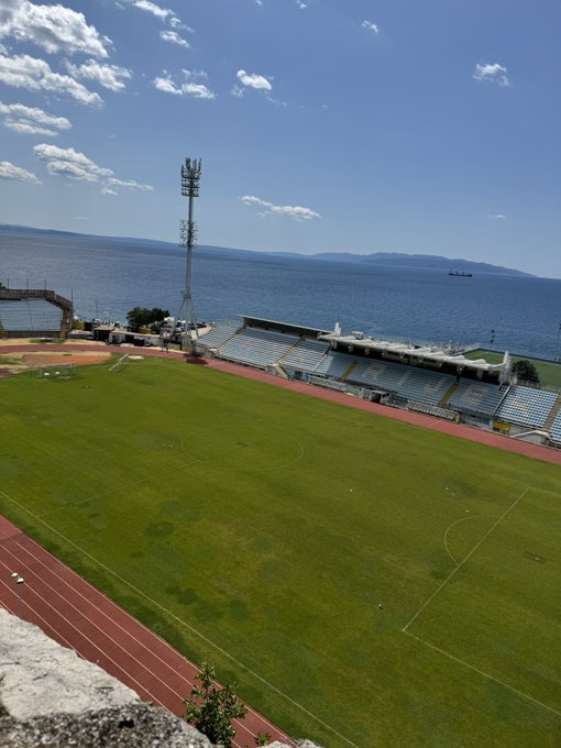 A coastal sports stadium featuring a green soccer field surrounded by a red running track, with bleacher seating and a communications tower overlooking the sea, juxtaposed against a promotional message for a crypto trading challenge. The serene, expansive stadium view contrasts with the high-stakes, promotional nature of the trading pitch.