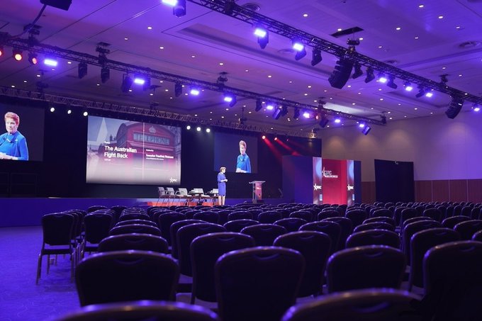 An empty conference hall with purple lighting shows a presenter on stage with screens displaying 'The Australian Fight Back' and a logo reading 'AU BARNES'.