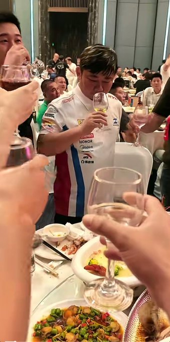 A crowded banquet scene where multiple people are raising wine glasses in a toast, with a person in the center wearing a shirt with sponsor logos including 'HONOR' and 'Snickers'.