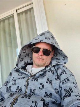 A man wearing sunglasses and a grey hoodie covered in a monogram pattern with the hood up.