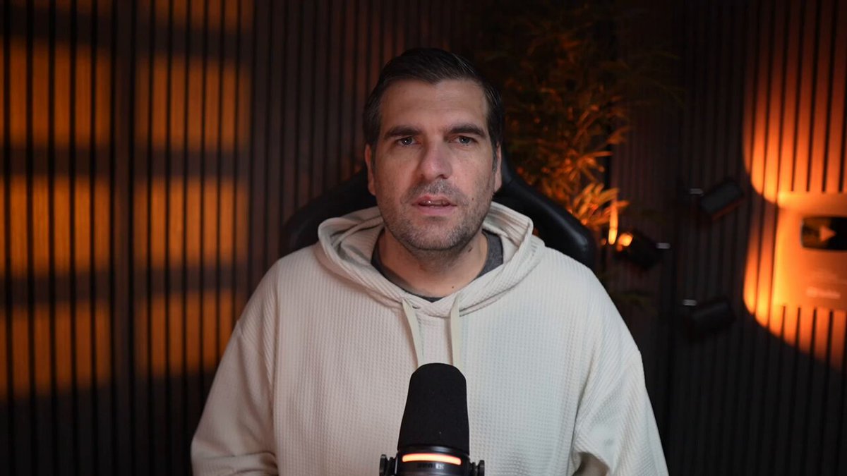 The image shows a man wearing a white hoodie, sitting behind a microphone with his hands on his knees. The background features a dark wall with a window that allows some light into the room. There are also two speakers visible in the scene. The man appears to be speaking or presenting something, possibly related to the post about Cardano's upcoming update and its impact on the network math.