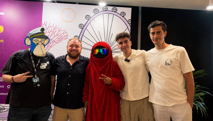 A group photo of five men posing in front of a backdrop, including someone in an ape mask and another in a red hooded robe with a reflective visor.