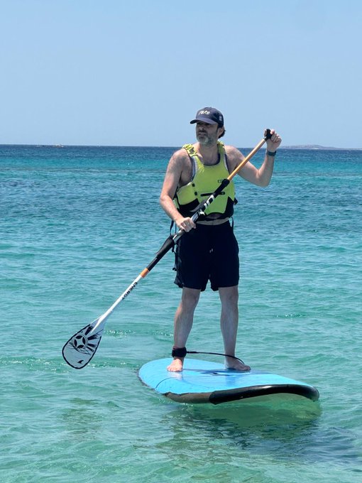 A man stands on a paddleboard in clear turquoise water, holding a paddle and wearing a yellow life vest and black cap.