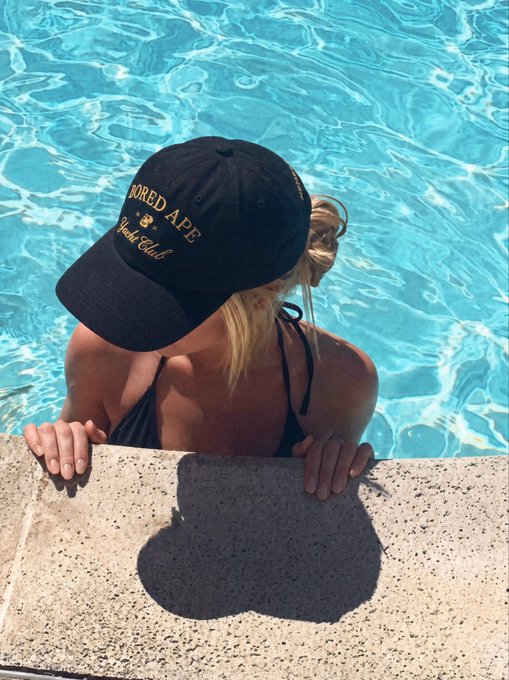 A woman in a black bikini is in a pool, leaning on the edge with a navy "Bored Ape Yacht Club" cap pulled down over her face.