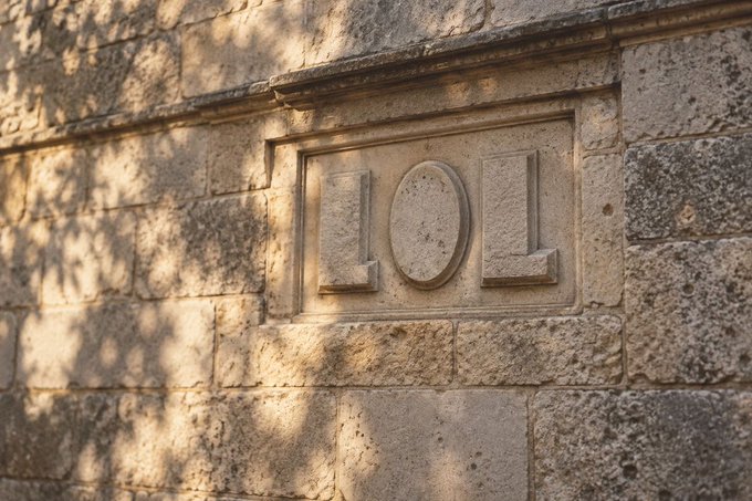 A stone wall with an embedded plaque displaying the carved letters "LOL", partially covered by the shadows of tree leaves.