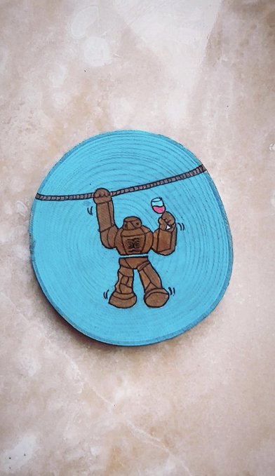 A small circular wooden slice painted turquoise blue, featuring a hand-drawn illustration of a brown robot flexing its muscles while holding a cup of red wine, displayed on a marble surface.
