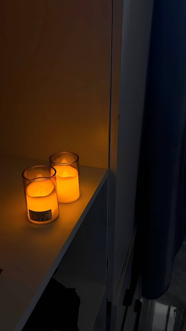 Two lit candles in glass jars sit on a white shelf in a dimly lit space.