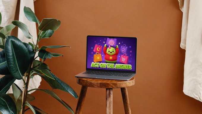 A laptop on a wooden stool displays a colorful graphic featuring a green character and shopping bags with the text "ACT ON THE ANSWER," set against an orange wall with a rubber plant.