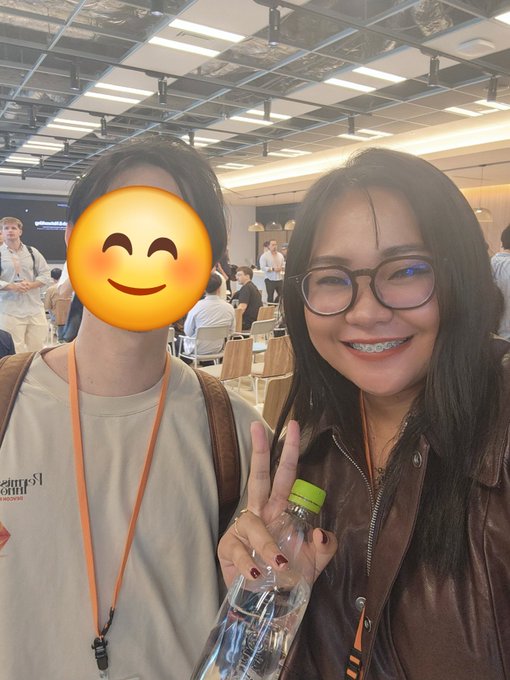 A photo of two young people at what appears to be a conference or event, with one person's face covered by a blushing smiley emoji, while the other young woman with glasses and braces smiles and makes a peace sign while holding a water bottle.