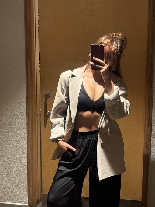 A mirror selfie captures a woman wearing an oversized white blazer over a black bra and black trousers, with the caption "I think it's quite explanatory" highlighting the bold styling choice of revealing a fitted top beneath the open, layered jacket.