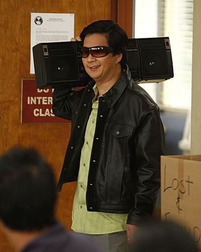 A scene from a TV show featuring Jackie Chan wearing sunglasses and a black leather jacket, smiling as he carries two large black speakers on his shoulders in front of a 'LOST & FOUND' sign.