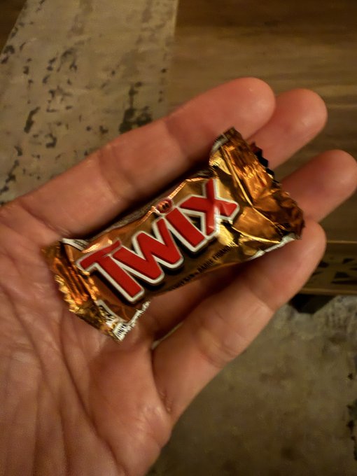 A hand holds a mini Twix candy bar in its gold foil wrapper, prominently displaying the red "TWIX" logo.