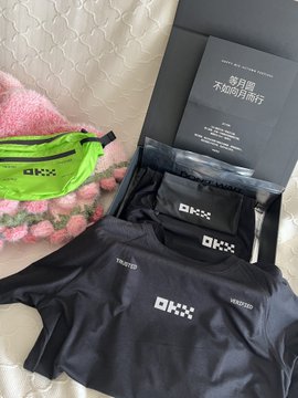 OKX merchandise including a black shirt, fanny pack, and booklet are laid out on a bed with a pink patterned pillow.