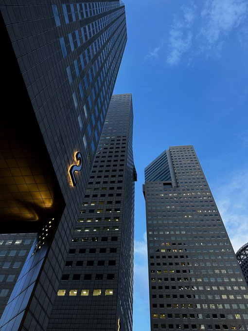 A dramatic upward view of towering modern skyscrapers against a deep blue sky, with a glowing "S" logo visible on the underside of a building's overhang—capturing the literal perspective of "looking up" at the impressive urban architecture.