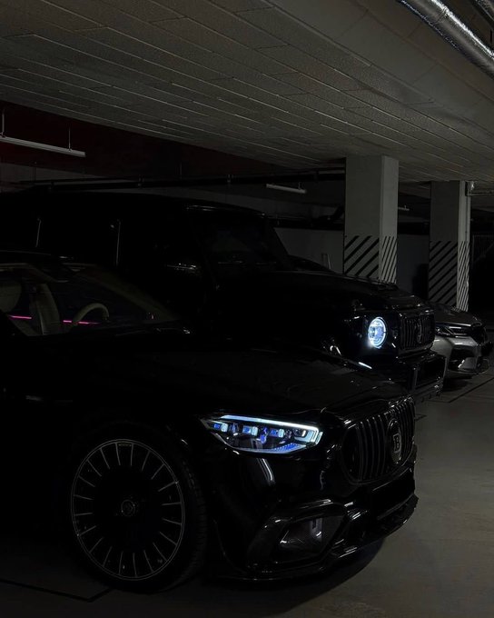 The image features a black sports car parked inside an underground garage with a white ceiling. The car has its headlights turned on, illuminating the scene. There are two other cars visible in the background, one of which appears to be a jeep or SUV. A person is also present in the image, standing near the second car.