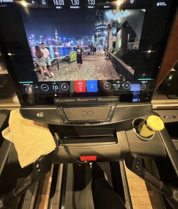 The image shows a person using an exercise machine at a gym, with their feet resting on a tread belt while they are working out. The machine displays a video screen showing people walking along a city street or a park, which suggests that this is likely a treadmill workout session. There is also a towel placed nearby the user's feet, indicating that they have been using it to wipe off sweat during their exercise routine.