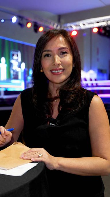 A portrait of a smiling woman with long dark hair wearing a black sleeveless top, seated at what appears to be a press or interview setting with stage lighting behind her.