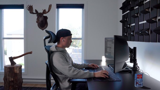 A person wearing a backwards cap and earbuds works at a computer desk in a modern office, with a decorative moose head mounted on the wall behind them.