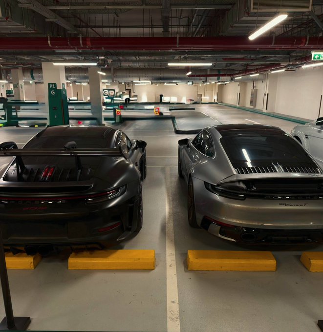 Two Porsche 911 GT3 cars—one black and one silver—are parked next to each other in a concrete underground parking garage, viewed from the rear.