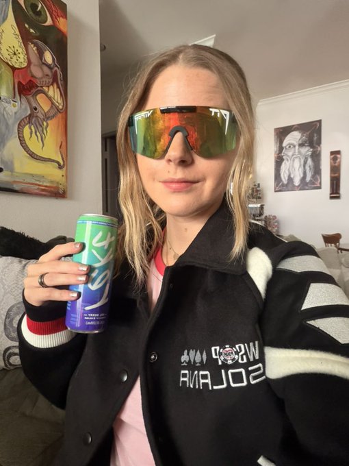 A young woman with blonde hair wears reflective orange-tinted sunglasses and a black letterman jacket reading 'WSP SOOLCRA' (appearing mirrored), while holding a green and blue 'Alani Nu' energy drink can in a living room with colorful artwork on the walls.