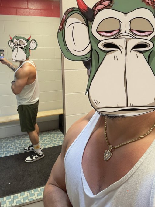 A man takes a mirror selfie in a locker room while wearing a white tank top, green shorts, a gold chain, and a digital gorilla mask filter on his face.