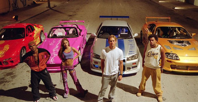 Four people stand in front of four brightly colored sports cars (red, pink, silver, and orange) in a nighttime setting, characteristic of the 2 Fast 2 Furious movie scene.