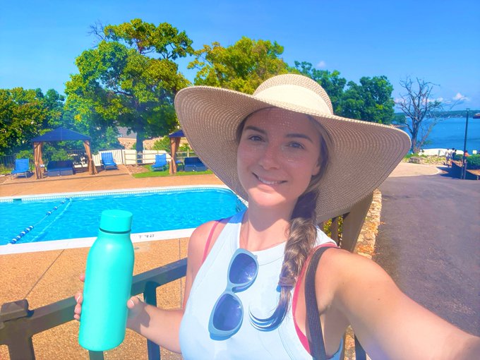 Selfie of a smiling woman wearing a wide-brimmed sun hat and white top, holding a teal water bottle near a pool with a lake in the background.