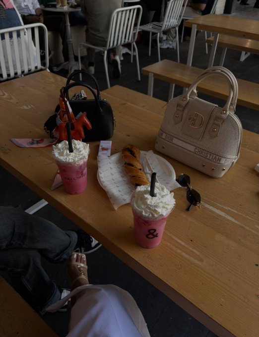 Two pink milkshakes topped with whipped cream sit on a wooden café table alongside a croissant, with two handbags (one black, one beige Christian Dior) in the background.