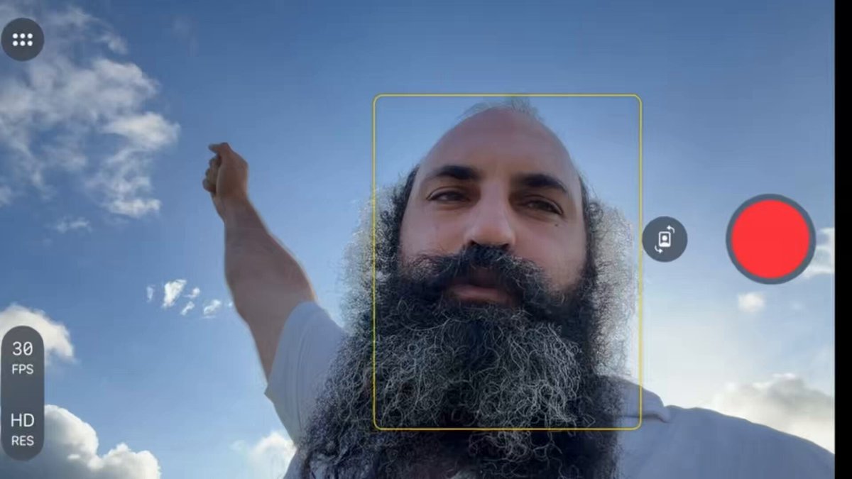 The image shows a man with a beard holding up his arm, possibly taking a selfie or recording something happening above him. The background of the photo features a blue sky with clouds, which adds an interesting visual element to the scene. There are also two red dots in the image, one located near the top left corner and another towards the bottom right corner. These red dots could be related to the post's content or might simply be part of the photo's composition. The man holding his arm up appears to be the main subject of the image, while the background elements provide context for the scene.